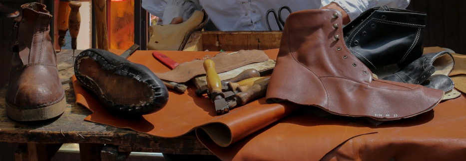 a series of leather shoes, uncut leather and leather-working tools laid out on a wooden workbench